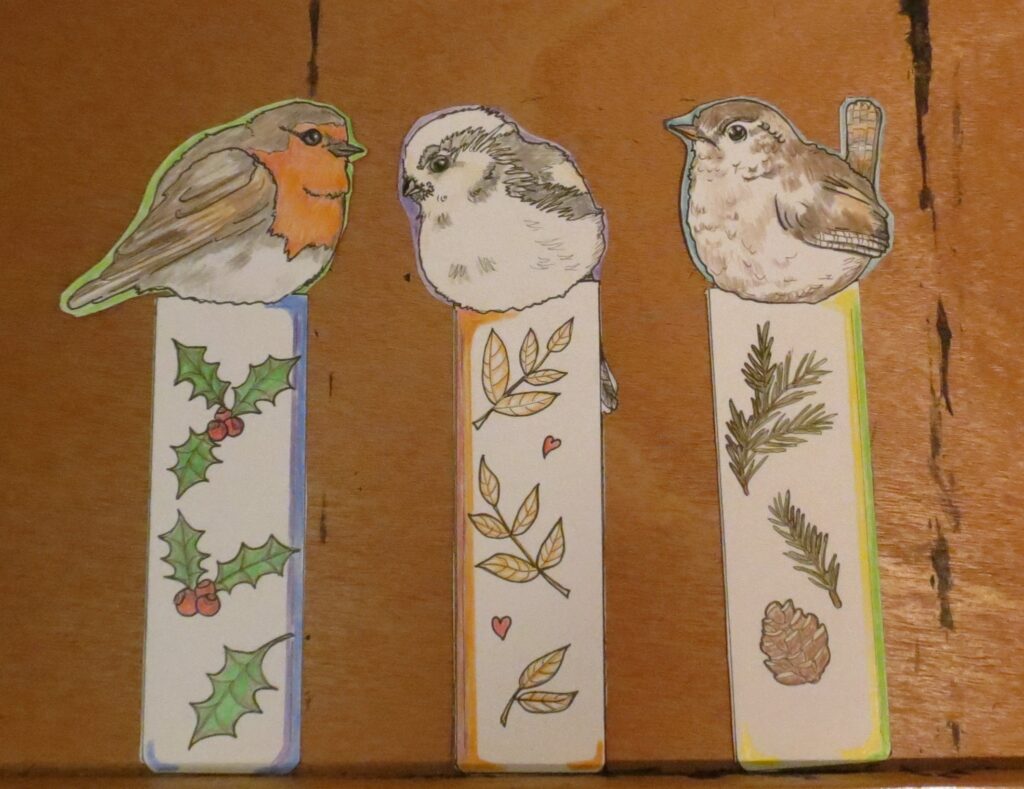 paper bookmarks showing 3 different cute birds. the first is a european robin with its orange chest and some thorny plants with red berries along the bookmark, the second is a longtailed tit, a white and grey little bird, the bookmark has brown leaves and heart, and the third is a wren, a little brown bird, the bookmark has a cone and needle twigs on it. the birds sit on top of the bookmark columns. the bookmarks are presented on a wooden easel