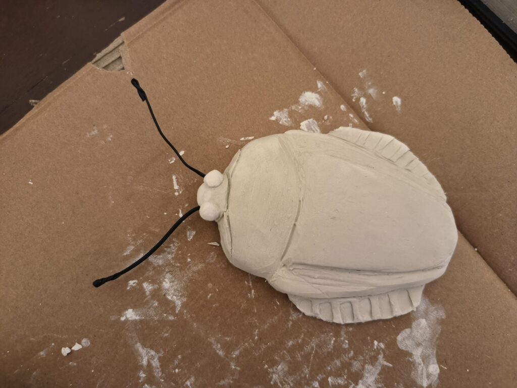 A raw sand colored ceramiclay sculpture of a flat shield bug. It doesn't have any color yet and is very fresh and still drying. Its antennae consists of two black metal cable binders.