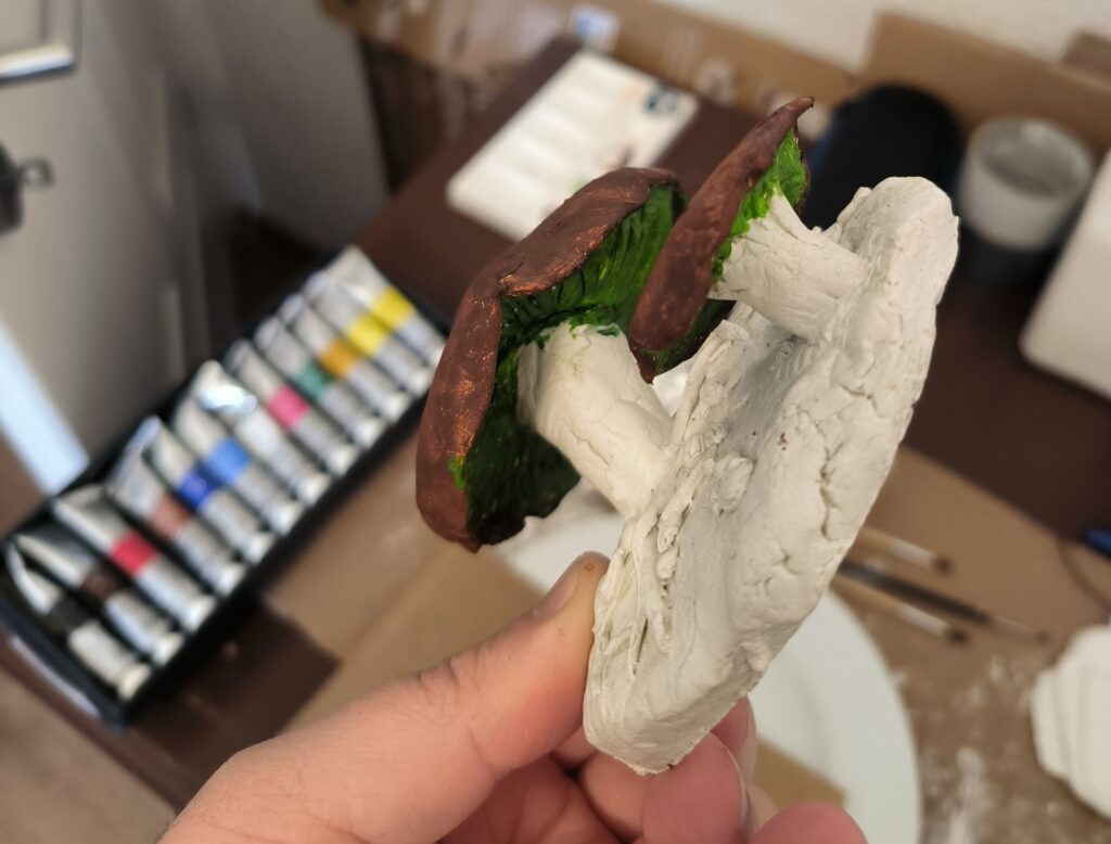 A crumbleing figure of two mushrooms. So far their hood is painted dark brown, even though a bit patchy, and the underside is green. But here and there there are green spots on the brown.